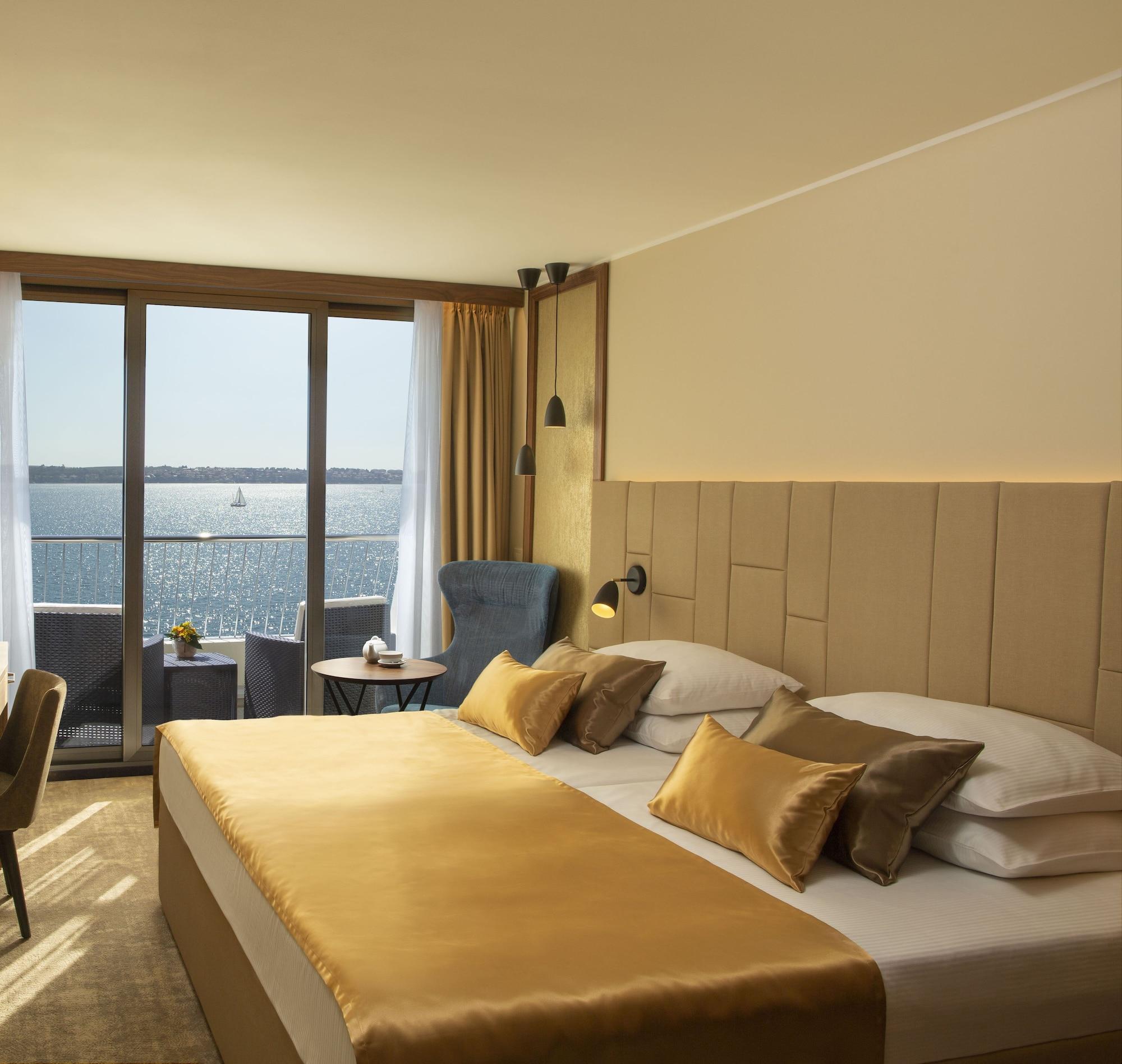 Grand Hotel Bernardin Portoroz Bagian luar foto The photo shows a cozy hotel room with a modern design. The room features a large bed with a beige and gold color scheme, complemented by decorative pillows. There is a seating area with a blue armchair and a small round table, positioned near a larg