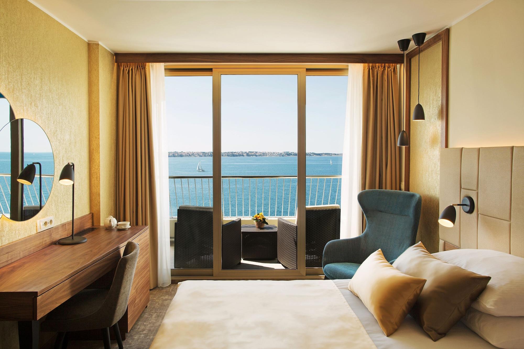 Grand Hotel Bernardin Portoroz Bagian luar foto The image shows a well-designed hotel room with a modern aesthetic. The room features a large bed with decorative pillows and a soft blanket. There's a stylish armchair in a blue hue next to a small table with two chairs, creating a cozy seating area