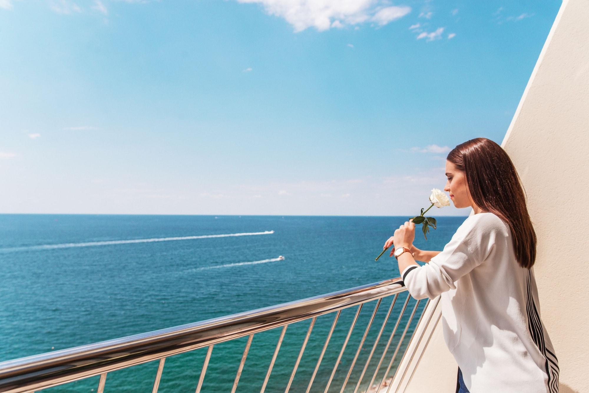 Grand Hotel Bernardin Portoroz Bagian luar foto The photo depicts a woman standing on a balcony overlooking the ocean. She holds a white rose and looks thoughtfully toward the water, which appears calm and blue. There are two boats visible on the surface of the sea, creating gentle waves. The sky 