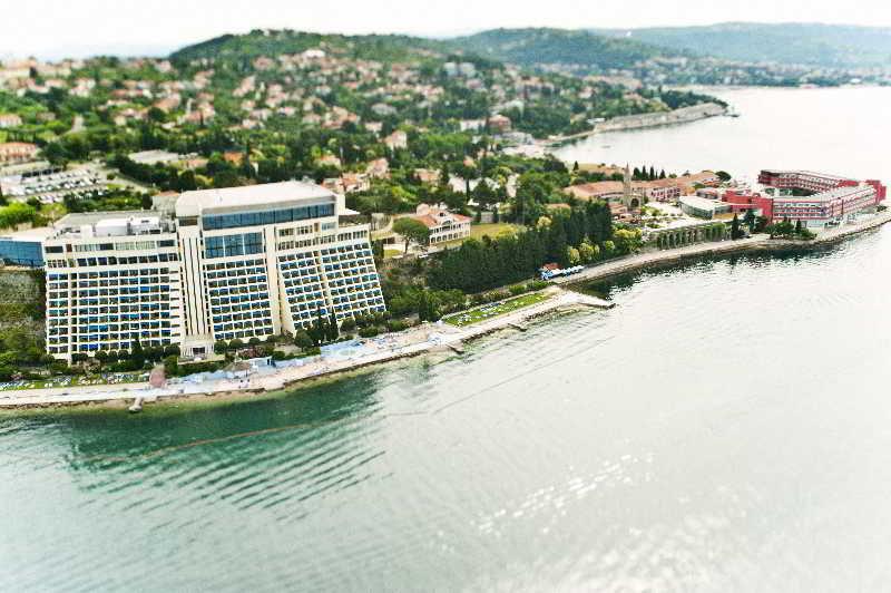 Grand Hotel Bernardin Portoroz Bagian luar foto The image shows an aerial view of a waterfront area. There is a large modern hotel or building with a lot of windows situated near the shore. Alongside the building, there is a beach area with what appears to be sun loungers. In the background, there