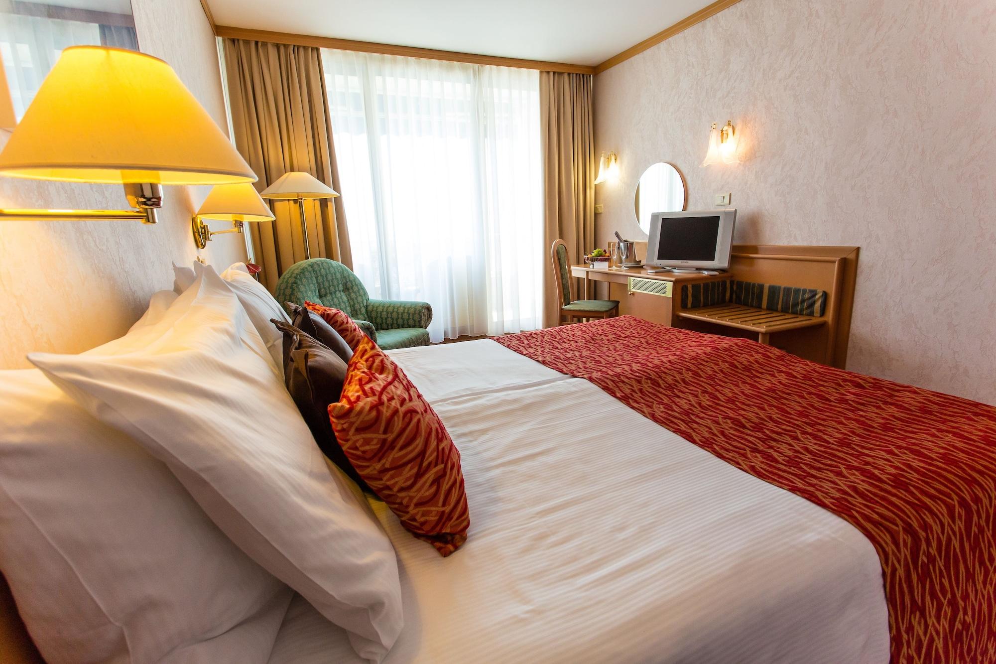 Grand Hotel Bernardin Portoroz Bagian luar foto The photo shows a hotel room featuring two beds, each with decorative pillows. There is a warm color scheme, highlighted by a red bedspread. To the side, a green armchair is visible, along with a desk that has a mirror and a television on it. Large w