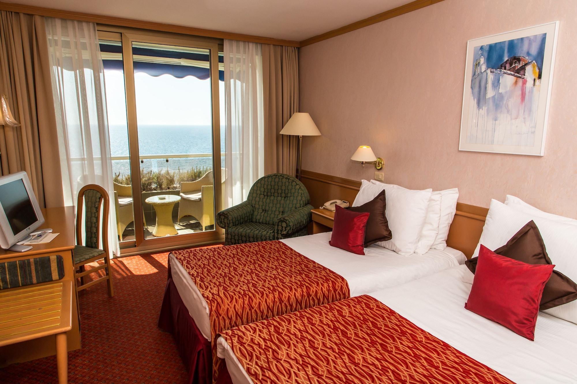 Grand Hotel Bernardin Portoroz Bagian luar foto The photo shows a hotel room featuring two twin beds with decorative red and white bedding. There is a small seating area with a green chair and a side table. The room has large windows that offer a view of the ocean, and a balcony with two chairs is
