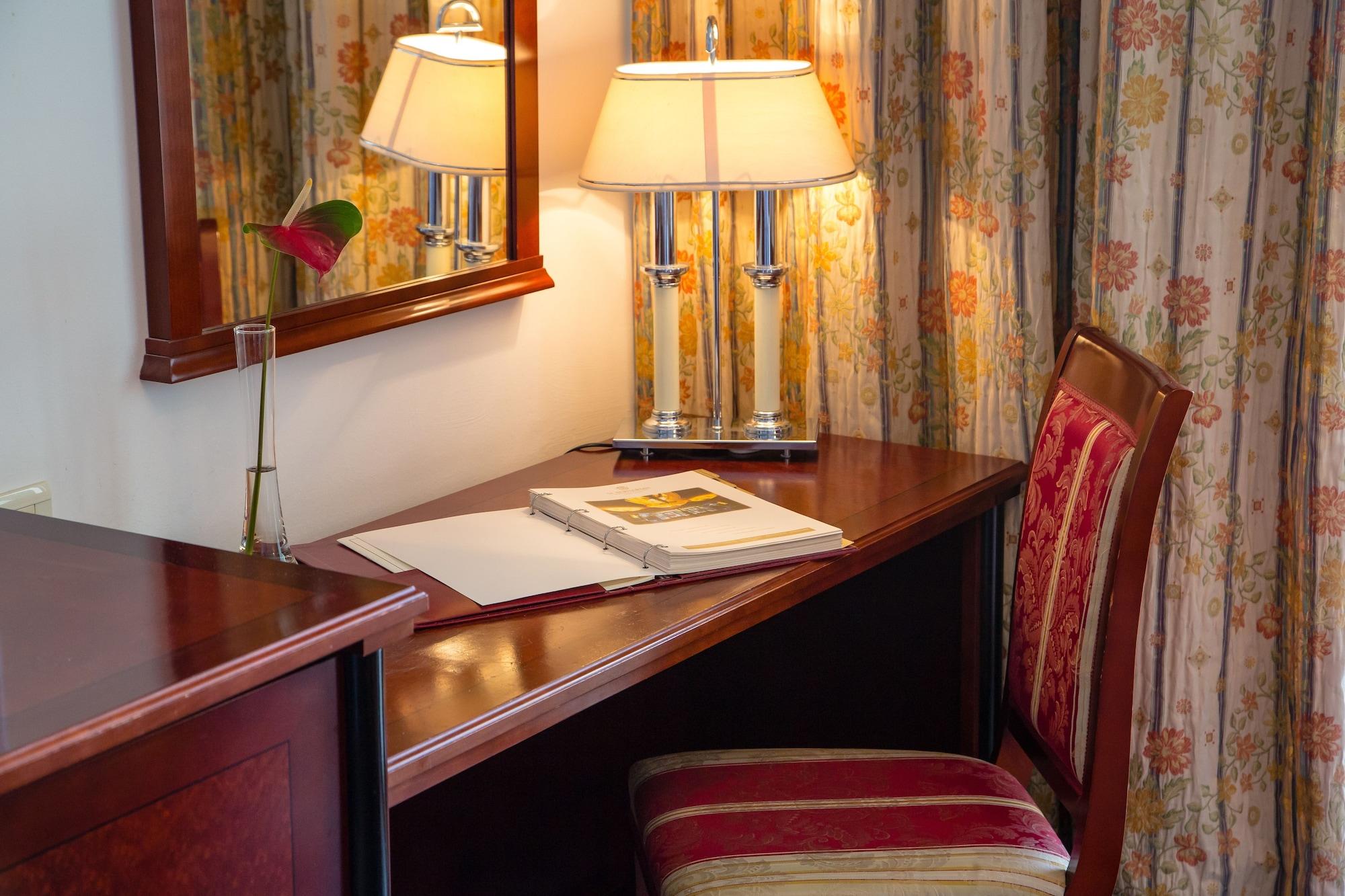 Grand Hotel Bernardin Portoroz Bagian luar foto The photo shows a cozy desk setup in a room. There's a wooden desk with a dark finish next to a comfortable chair with a striped cushion. On the desk, a few items are placed, including an open book or notebook and a small vase with a flower. Above th