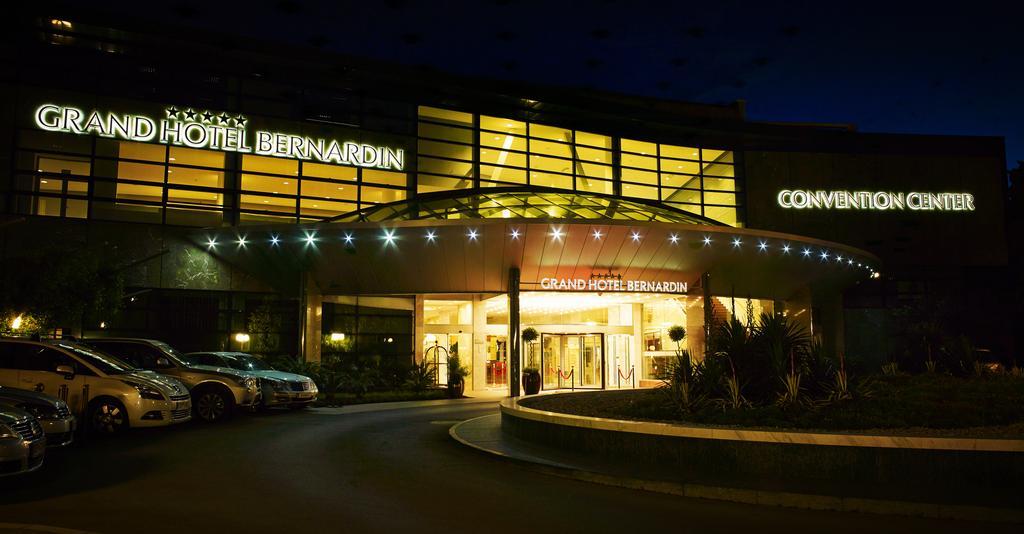 Grand Hotel Bernardin Portoroz Bagian luar foto The photo shows a building, identified as the Grand Hotel Bernardin, illuminated at night. The entrance features a curved awning with decorative lights, creating a welcoming atmosphere. The front area has several parked cars and large glass windows, 