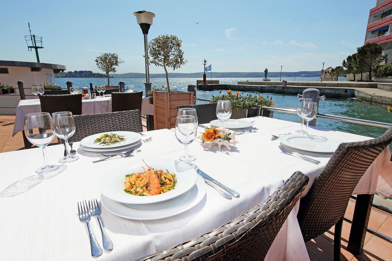 Grand Hotel Bernardin Portoroz Bagian luar foto The photo shows a picturesque outdoor dining setup by a waterfront. There are several tables with elegant white tablecloths, each set with plates, cutlery, and wine glasses. In the foreground, a plate of pasta with salmon is prominently displayed alo