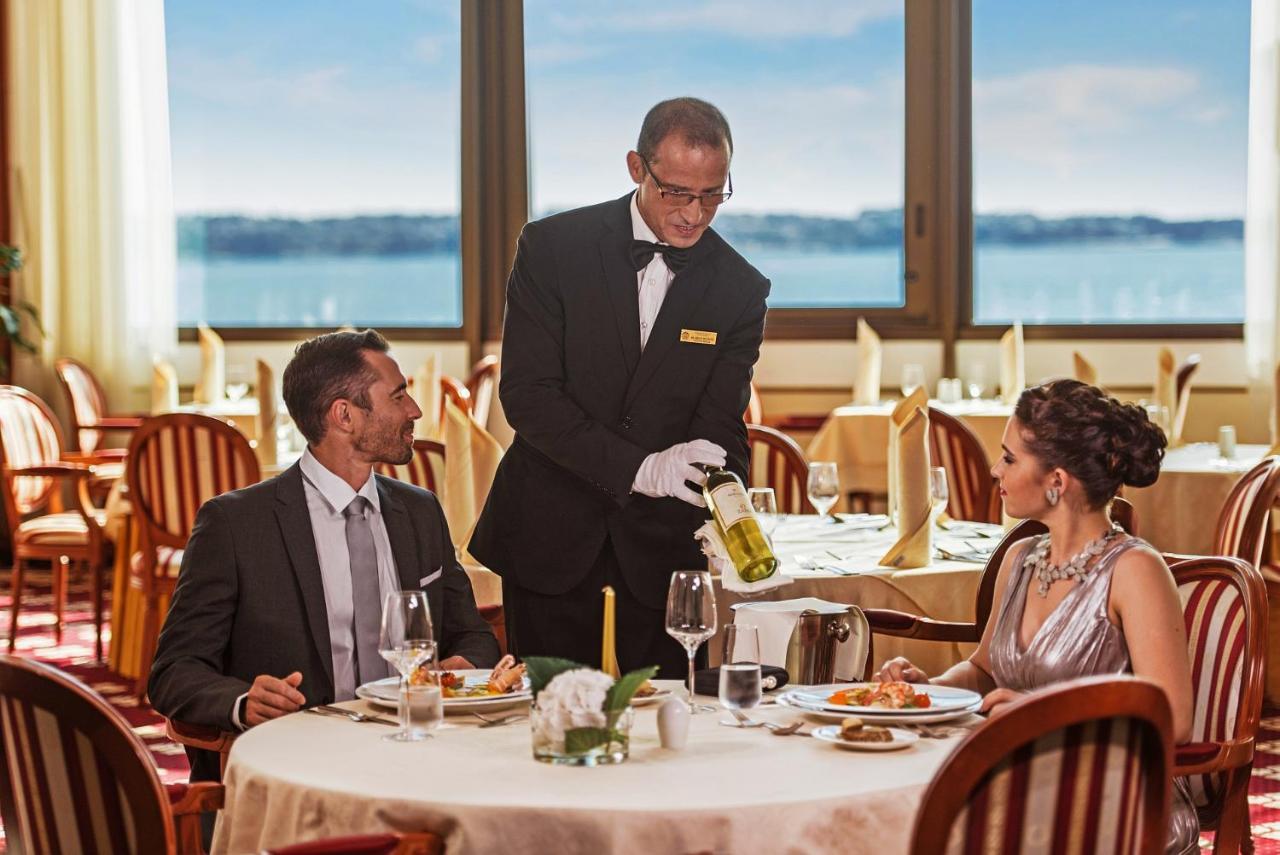 Grand Hotel Bernardin Portoroz Bagian luar foto The image shows a fine dining setting in a restaurant. In the foreground, a well-dressed couple is seated at an elegantly set table. The man is looking towards the waiter, who is pouring a drink from a bottle into a wine glass. The woman is sitting b