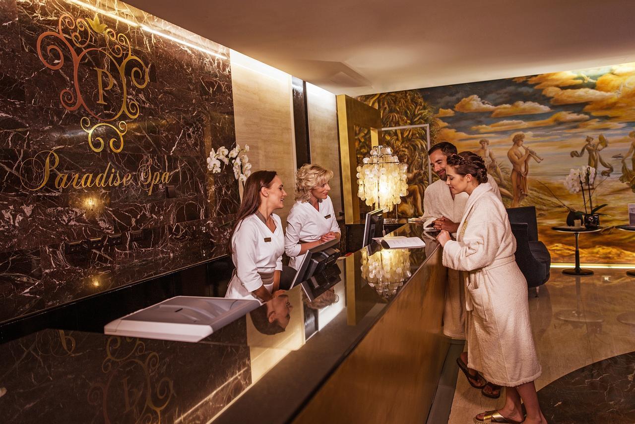 Grand Hotel Bernardin Portoroz Bagian luar foto The photo shows a luxurious spa reception area. There are three staff members behind a sleek, dark marble reception desk, wearing white robes. Two guests, also in robes, are interacting with the staff. The background features an elegant mural and sty