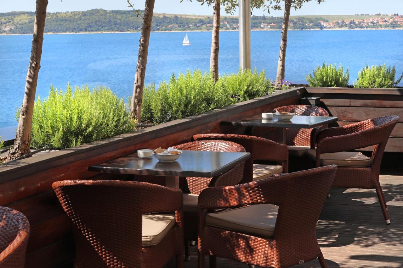 Grand Hotel Bernardin Portoroz Bagian luar foto The photo shows an outdoor seating area of a restaurant or café with several wicker chairs and tables. The setting is surrounded by green plants and has a view of a calm body of water, likely a lake or sea, in the background. In the distance, there's